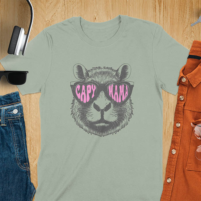 A t-shirt featuring a capybara face with pink heart-shaped sunglasses that spell CAPY MAMA. Below the capybara, Meagan is written in a pink, script font. This vintage mama shirt design is a retro mom tee-shirt perfect for mama mom life. A trendy mama tee for an expecting mom or pregnant mom gift, ideal for a pregnancy announce or pregnancy reveal. A great mom gift hers or mother days gift, or a new mom gift for a gift for wife. Personalization is available by changing the Meagan name.