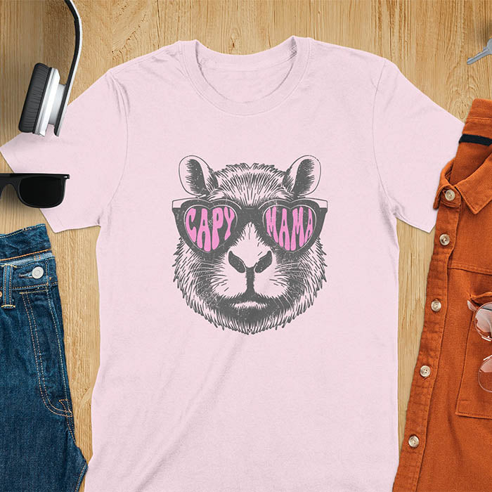 A t-shirt featuring a capybara face with pink heart-shaped sunglasses that spell CAPY MAMA. Below the capybara, Meagan is written in a pink, script font. This vintage mama shirt design is a retro mom tee-shirt perfect for mama mom life. A trendy mama tee for an expecting mom or pregnant mom gift, ideal for a pregnancy announce or pregnancy reveal. A great mom gift hers or mother days gift, or a new mom gift for a gift for wife. Personalization is available by changing the Meagan name.