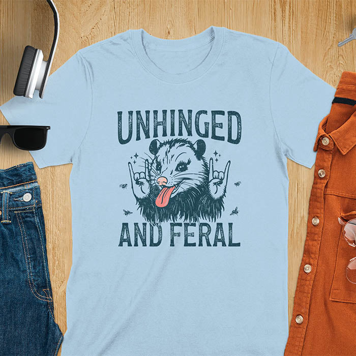 A t-shirt featuring a opossum with its tongue sticking out, throwing rock-on hand gestures. Small flies surround the opossum. Text: UNHINGED above and AND FERAL below in a distressed font. This unhinged shirt features a funny opossum design, a weird meme shirt or weird t shirt for unhinged gifts. A sarcasm shirt with a sassy shirt attitude, a memeing shirt or genz meme for an animal person gift.