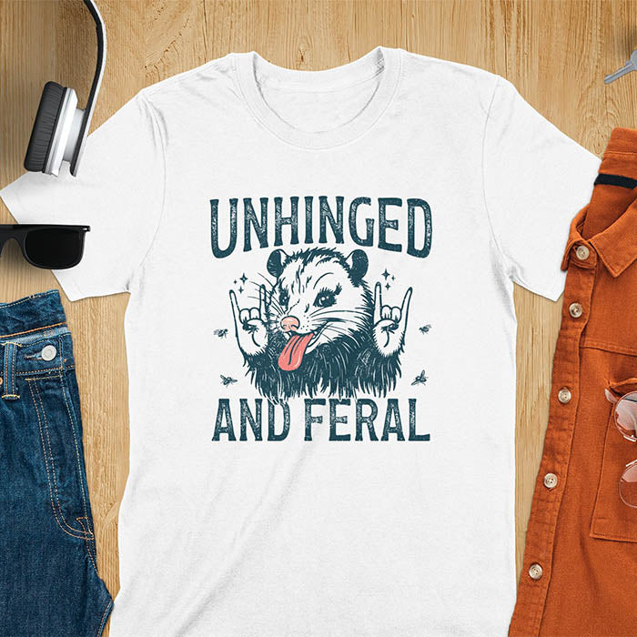 A t-shirt featuring a opossum with its tongue sticking out, throwing rock-on hand gestures. Small flies surround the opossum. Text: UNHINGED above and AND FERAL below in a distressed font. This unhinged shirt features a funny opossum design, a weird meme shirt or weird t shirt for unhinged gifts. A sarcasm shirt with a sassy shirt attitude, a memeing shirt or genz meme for an animal person gift.