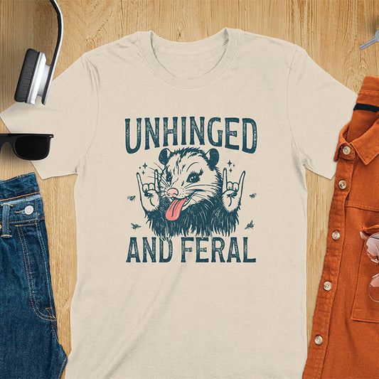 A t-shirt featuring a opossum with its tongue sticking out, throwing rock-on hand gestures. Small flies surround the opossum. Text: UNHINGED above and AND FERAL below in a distressed font. This unhinged shirt features a funny opossum design, a weird meme shirt or weird t shirt for unhinged gifts. A sarcasm shirt with a sassy shirt attitude, a memeing shirt or genz meme for an animal person gift.