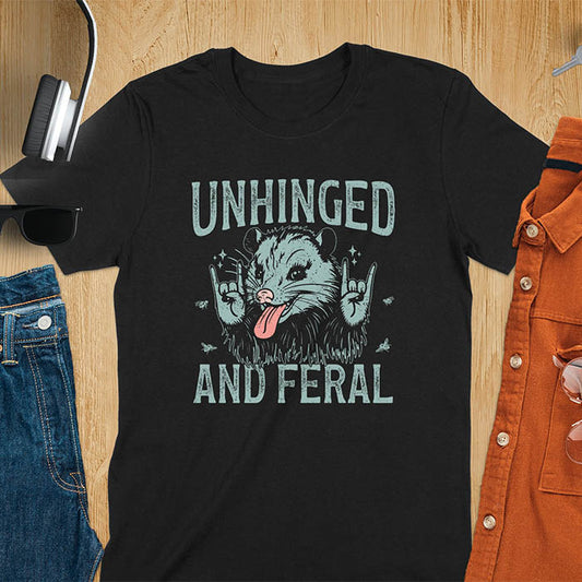 A t-shirt featuring a opossum with its tongue sticking out, throwing rock-on hand gestures. Small flies surround the opossum. Text: UNHINGED above and AND FERAL below in a distressed font. This unhinged shirt features a funny opossum design, a weird meme shirt or weird t shirt for unhinged gifts. A sarcasm shirt with a sassy shirt attitude, a memeing shirt or genz meme for an animal person gift.