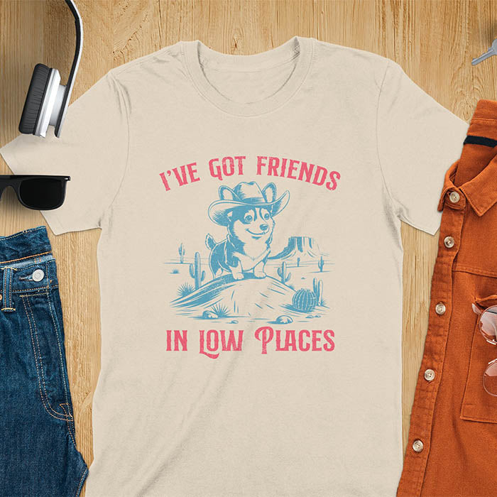 A t-shirt with a corgi wearing a cowboy hat sitting in a desert scene with cacti. Text: "I've Got FRIENDS" above and "IN LOW PLACES" below. This retro dog shirt is a corgi shirt and funny dog shirt, a vintage dog tee. A cowboy tshirt for a dog dad shirt and pet owner gift. An animal person gift with cuteness puppy, a meme graphic tee, a mom gift option.