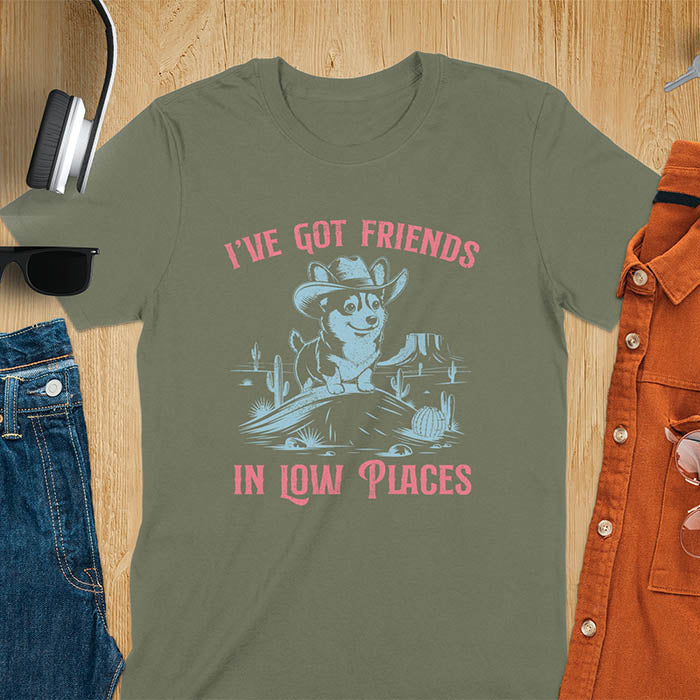 A t-shirt with a corgi wearing a cowboy hat sitting in a desert scene with cacti. Text: "I've Got FRIENDS" above and "IN LOW PLACES" below. This retro dog shirt is a corgi shirt and funny dog shirt, a vintage dog tee. A cowboy tshirt for a dog dad shirt and pet owner gift. An animal person gift with cuteness puppy, a meme graphic tee, a mom gift option.