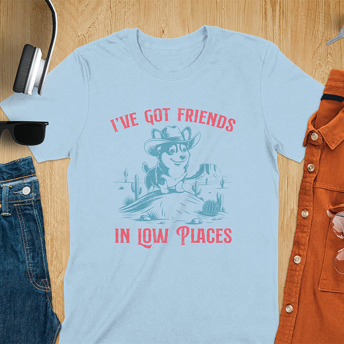 A t-shirt with a corgi wearing a cowboy hat sitting in a desert scene with cacti. Text: "I've Got FRIENDS" above and "IN LOW PLACES" below. This retro dog shirt is a corgi shirt and funny dog shirt, a vintage dog tee. A cowboy tshirt for a dog dad shirt and pet owner gift. An animal person gift with cuteness puppy, a meme graphic tee, a mom gift option.