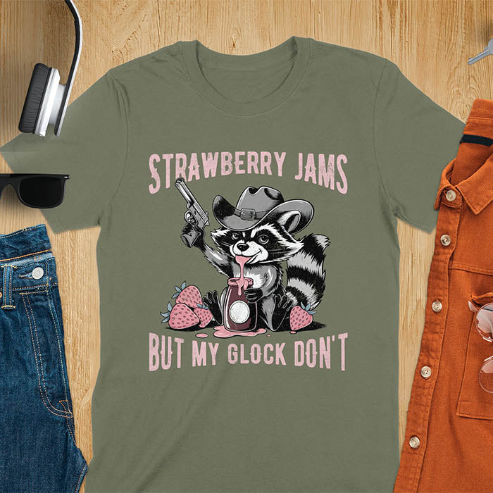 Funny raccoon t-shirt with a quirky raccoon in a cowboy hat eating strawberry jam while holding a gun. Pastel pink jam and strawberries pop against a distressed monochrome design. Text says Strawberry Jams above and But My Glock Don’t below.