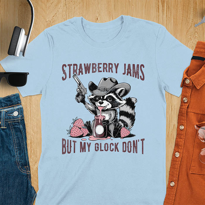 Funny raccoon t-shirt with a quirky raccoon in a cowboy hat eating strawberry jam while holding a gun. Pastel pink jam and strawberries pop against a distressed monochrome design. Text says Strawberry Jams above and But My Glock Don’t below.