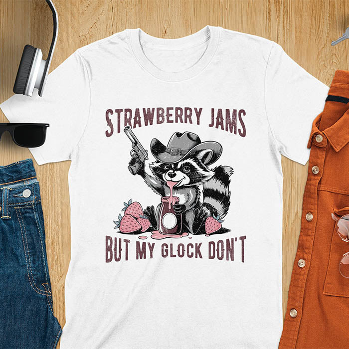 Funny raccoon t-shirt with a quirky raccoon in a cowboy hat eating strawberry jam while holding a gun. Pastel pink jam and strawberries pop against a distressed monochrome design. Text says Strawberry Jams above and But My Glock Don’t below.