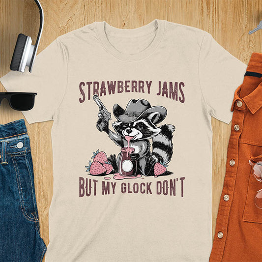 Funny raccoon t-shirt with a quirky raccoon in a cowboy hat eating strawberry jam while holding a gun. Pastel pink jam and strawberries pop against a distressed monochrome design. Text says Strawberry Jams above and But My Glock Don’t below.