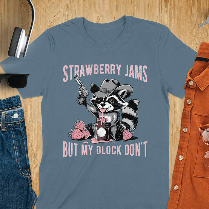 Funny raccoon t-shirt with a quirky raccoon in a cowboy hat eating strawberry jam while holding a gun. Pastel pink jam and strawberries pop against a distressed monochrome design. Text says Strawberry Jams above and But My Glock Don’t below.