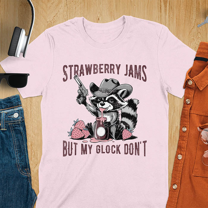 Funny raccoon t-shirt with a quirky raccoon in a cowboy hat eating strawberry jam while holding a gun. Pastel pink jam and strawberries pop against a distressed monochrome design. Text says Strawberry Jams above and But My Glock Don’t below.