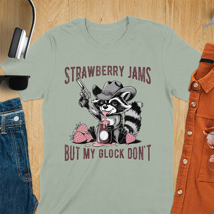Funny raccoon t-shirt with a quirky raccoon in a cowboy hat eating strawberry jam while holding a gun. Pastel pink jam and strawberries pop against a distressed monochrome design. Text says Strawberry Jams above and But My Glock Don’t below.