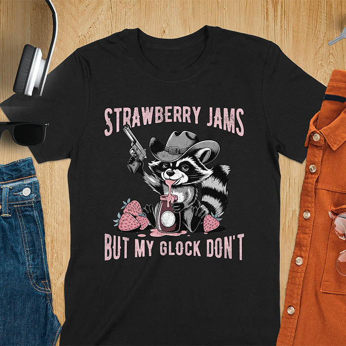 Funny raccoon t-shirt with a quirky raccoon in a cowboy hat eating strawberry jam while holding a gun. Pastel pink jam and strawberries pop against a distressed monochrome design. Text says Strawberry Jams above and But My Glock Don’t below.