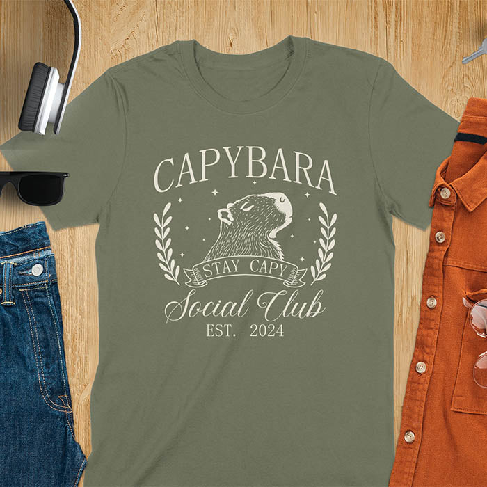 Green t-shirt with capybara design and text on a wooden surface with accessories. Stay capy slogan.