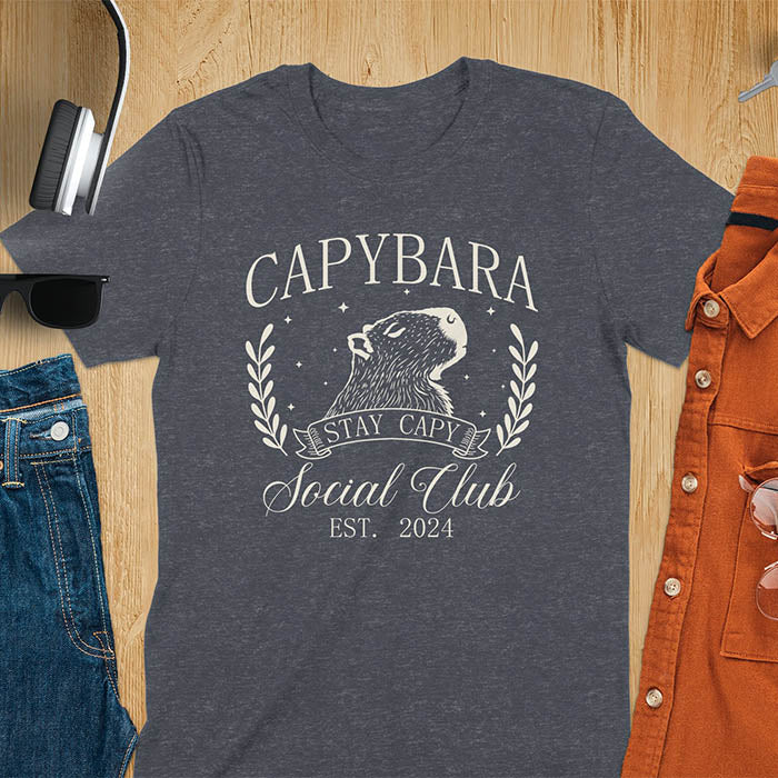 Gray t-shirt with 'Capybara Stay Capy Social Club' design on a wooden surface with accessories.