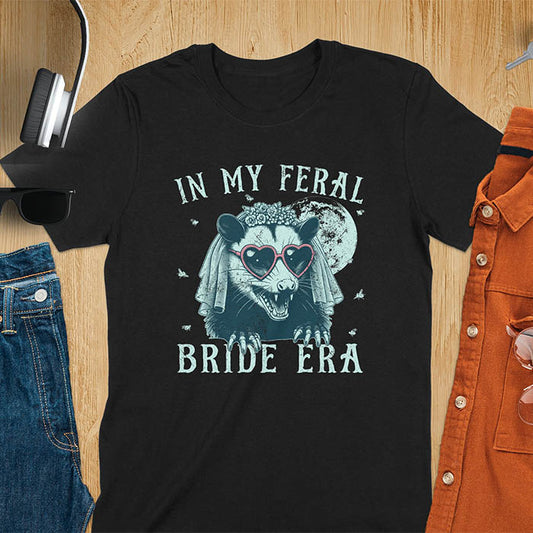 A t-shirt with IN MY FERAL BRIDE ERA in gothic font. Features an opossum bride with veil, claws, flies around her, and full moon in the background. Vintage-style with worn-out texture.
