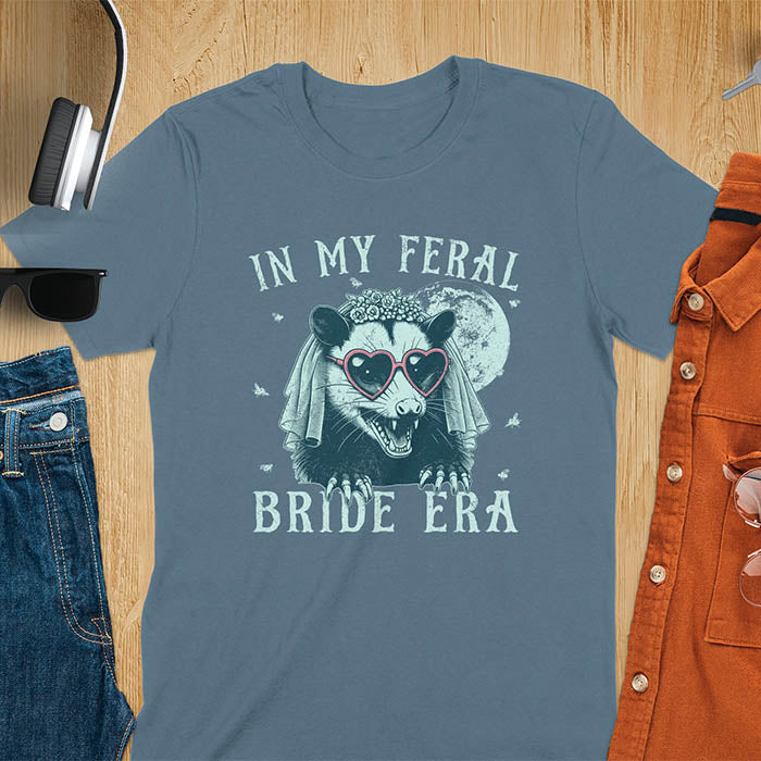 A t-shirt with IN MY FERAL BRIDE ERA in gothic font. Features an opossum bride with veil, claws, flies around her, and full moon in the background. Vintage-style with worn-out texture.
