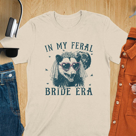 A t-shirt with IN MY FERAL BRIDE ERA in gothic font. Features an opossum bride with veil, claws, flies around her, and full moon in the background. Vintage-style with worn-out texture.