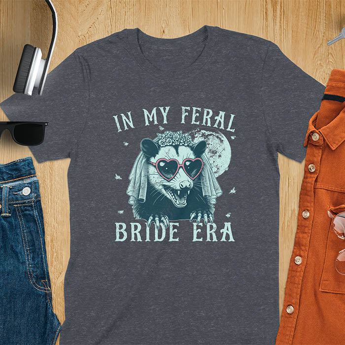 A t-shirt with IN MY FERAL BRIDE ERA in gothic font. Features an opossum bride with veil, claws, flies around her, and full moon in the background. Vintage-style with worn-out texture.