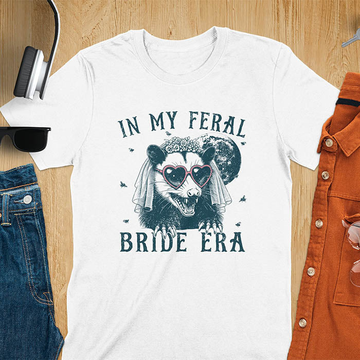 A t-shirt with IN MY FERAL BRIDE ERA in gothic font. Features an opossum bride with veil, claws, flies around her, and full moon in the background. Vintage-style with worn-out texture.