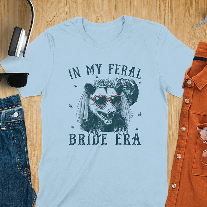 A t-shirt with IN MY FERAL BRIDE ERA in gothic font. Features an opossum bride with veil, claws, flies around her, and full moon in the background. Vintage-style with worn-out texture.