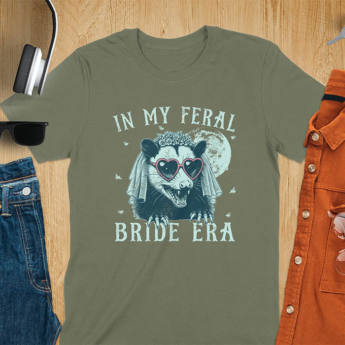 A t-shirt with IN MY FERAL BRIDE ERA in gothic font. Features an opossum bride with veil, claws, flies around her, and full moon in the background. Vintage-style with worn-out texture.