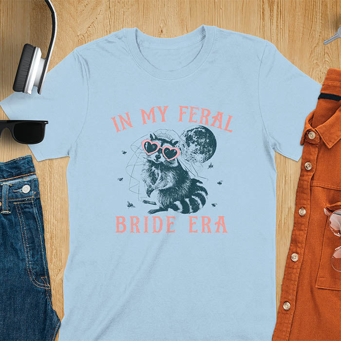 A raccoon wearing heart-shaped sunglasses and a bride's veil, with the moon in the background. Text reads In My Feral Bride Era.
