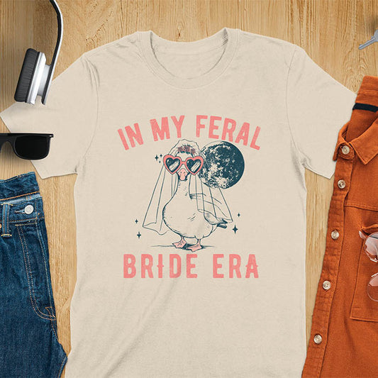 A goose standing, wearing heart-shaped sunglasses and a bride's veil. Moon in the upper right background. Retro-style text reads In My Feral on top Bride Era at bottom. Whimsical image.