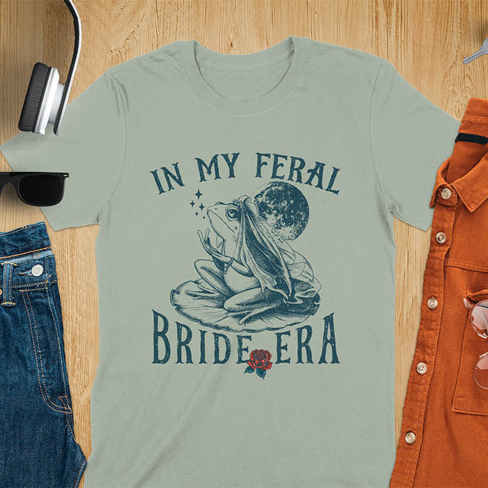 A vintage-style frog wearing a bride veil, croaking to the sky from a lily pad with a moon in the background. Text reads In My Feral Bride Era.