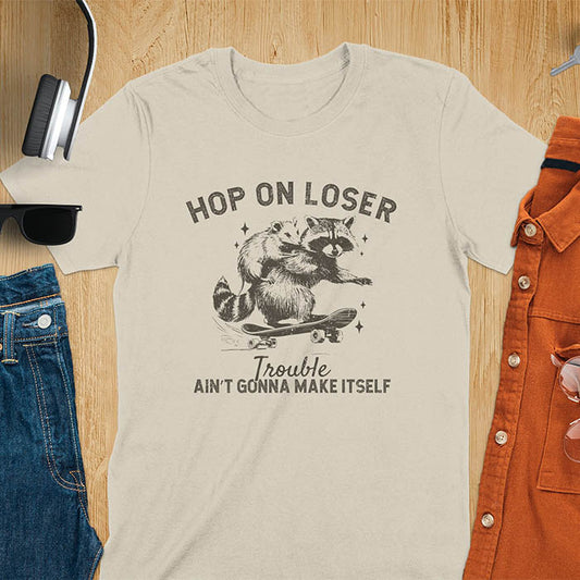 A raccoon riding a skateboard with an opossum on its back. Text reads Hop On Loser and Trouble Ain't Gonna Make Itself.