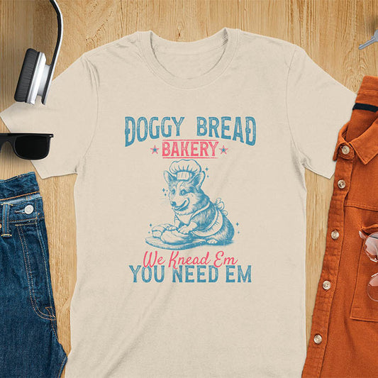 A t-shirt with a corgi wearing a chef's hat and apron, kneading dough. Text: "DOGGY BREAD BAKERY" above and "We Knead Em YOU NEED EM" below. This funny baking shirt is a funny meme shirt and funny dog shirt showing corgi cuteness, a great dog lover gift and Gift for dog mom. An animal lovers tshirt and Dog person tee, a pet dad t-shirt or pet dog owner gift. One of the funny bakers gifts, a Gift for mom.