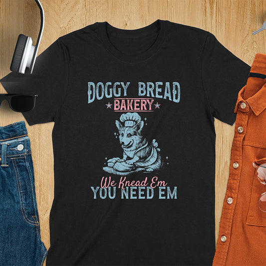 A t-shirt with a corgi wearing a chef's hat and apron, kneading dough. Text: "DOGGY BREAD BAKERY" above and "We Knead Em YOU NEED EM" below. This funny baking shirt is a funny meme shirt and funny dog shirt showing corgi cuteness, a great dog lover gift and Gift for dog mom. An animal lovers tshirt and Dog person tee, a pet dad t-shirt or pet dog owner gift. One of the funny bakers gifts, a Gift for mom.