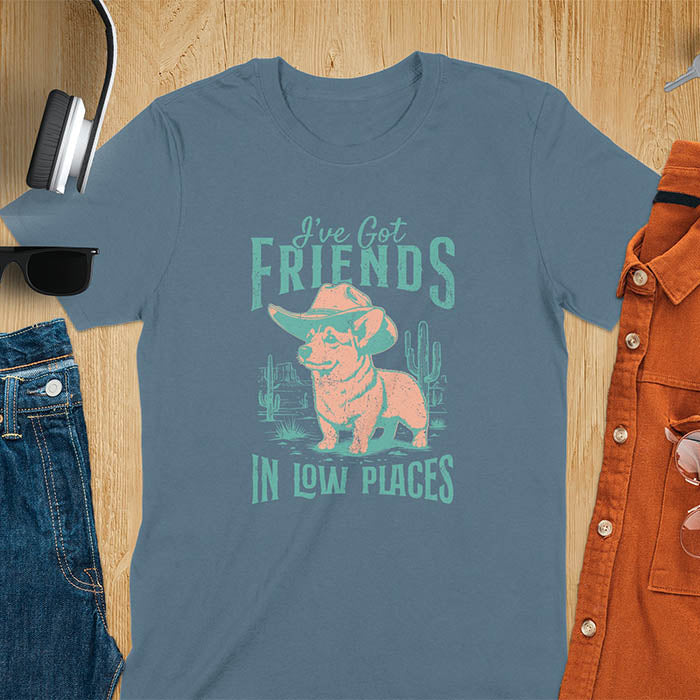A t-shirt with a corgi wearing a cowboy hat standing in a desert scene with cacti. Text: "I've Got FRIENDS" above and "IN LOW PLACES" below. This retro dog shirt is a corgi shirt and funny dog shirt, a vintage dog tee. A cowboy tshirt for a dog dad shirt and pet owner gift. An animal person gift with cuteness puppy, a meme graphic tee, a mom gift option.