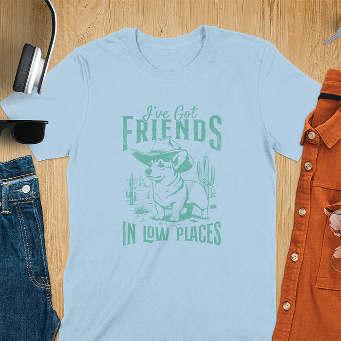 A t-shirt with a corgi wearing a cowboy hat standing in a desert scene with cacti. Text: "I've Got FRIENDS" above and "IN LOW PLACES" below. This retro dog shirt is a corgi shirt and funny dog shirt, a vintage dog tee. A cowboy tshirt for a dog dad shirt and pet owner gift. An animal person gift with cuteness puppy, a meme graphic tee, a mom gift option.