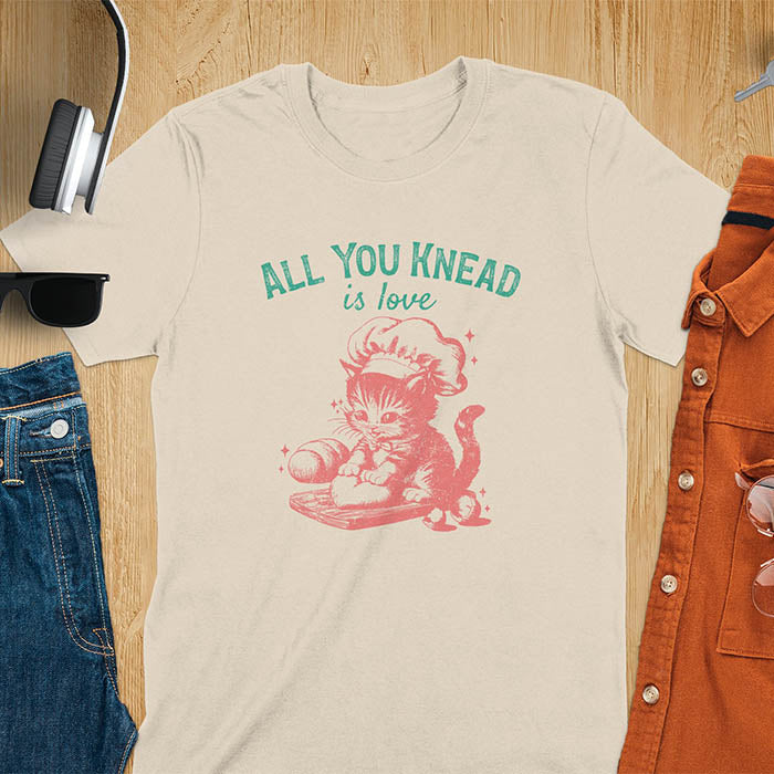 A t-shirt with a kitten wearing a chef's hat, kneading dough. Text: "ALL YOU knead" above and "is love" below. This Funny baking shirt is a Funny meme shirt showing kitten cuteness, a great cat lover gift and mom gift. An Animal lovers tshirt and cat person tee, a Pet cat owner gift. One of the Funny bakers gifts, a baker shirt gift, and a Funny cat t shirt.