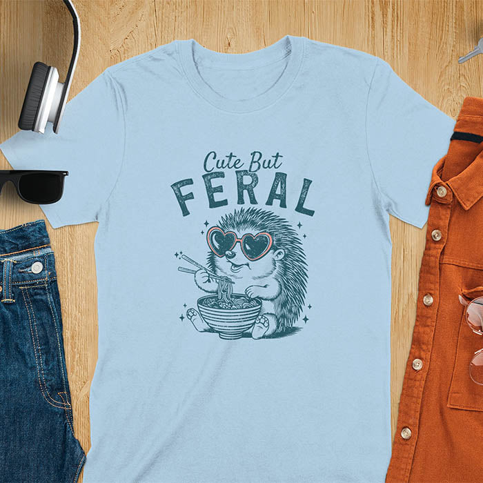 A t-shirt with a hedgehog wearing heart sunglasses and eating ramen with chopsticks. Text: "Cute But" above and "FERAL" below. This kawaiiness cuteness shows hedgehog cuteness in a weird funny shirt while ramen eating, a funny animal meme. A meme graphic tee and funny unhinge shirt, a great animal person gift and pet owner gift. A gen z meme with retro aesthetics tee vibes, a gift for mom or hers option, not quite wild safari animals.