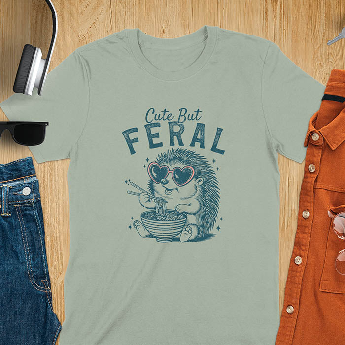 A t-shirt with a hedgehog wearing heart sunglasses and eating ramen with chopsticks. Text: "Cute But" above and "FERAL" below. This kawaiiness cuteness shows hedgehog cuteness in a weird funny shirt while ramen eating, a funny animal meme. A meme graphic tee and funny unhinge shirt, a great animal person gift and pet owner gift. A gen z meme with retro aesthetics tee vibes, a gift for mom or hers option, not quite wild safari animals.