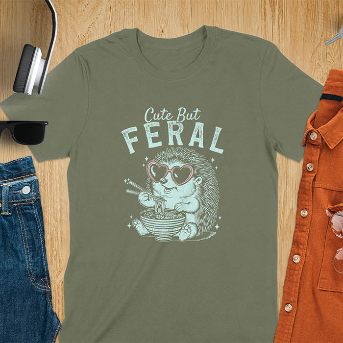 A t-shirt with a hedgehog wearing heart sunglasses and eating ramen with chopsticks. Text: "Cute But" above and "FERAL" below. This kawaiiness cuteness shows hedgehog cuteness in a weird funny shirt while ramen eating, a funny animal meme. A meme graphic tee and funny unhinge shirt, a great animal person gift and pet owner gift. A gen z meme with retro aesthetics tee vibes, a gift for mom or hers option, not quite wild safari animals.