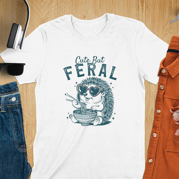 A t-shirt with a hedgehog wearing heart sunglasses and eating ramen with chopsticks. Text: "Cute But" above and "FERAL" below. This kawaiiness cuteness shows hedgehog cuteness in a weird funny shirt while ramen eating, a funny animal meme. A meme graphic tee and funny unhinge shirt, a great animal person gift and pet owner gift. A gen z meme with retro aesthetics tee vibes, a gift for mom or hers option, not quite wild safari animals.