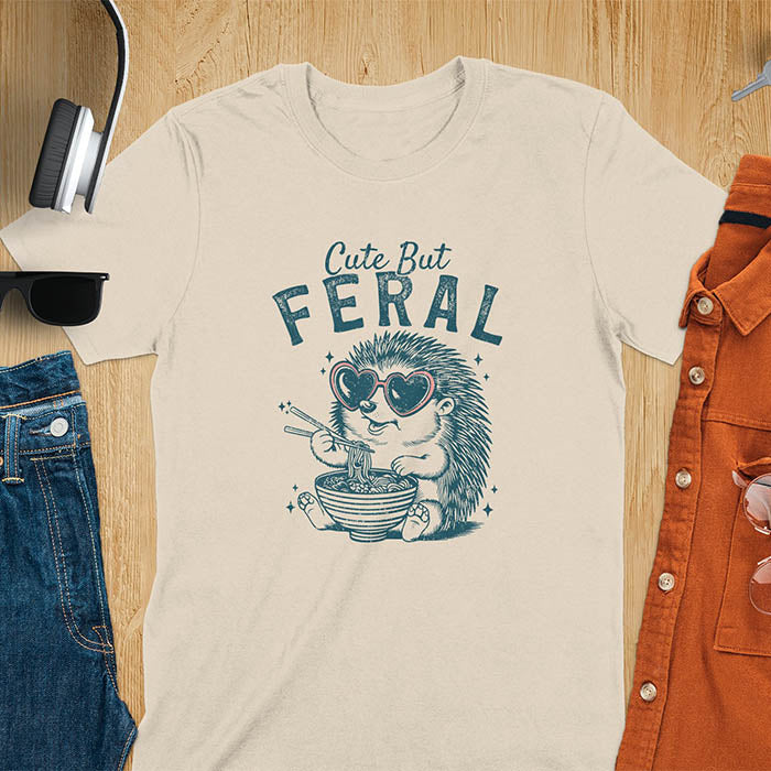 A t-shirt with a hedgehog wearing heart sunglasses and eating ramen with chopsticks. Text: "Cute But" above and "FERAL" below. This kawaiiness cuteness shows hedgehog cuteness in a weird funny shirt while ramen eating, a funny animal meme. A meme graphic tee and funny unhinge shirt, a great animal person gift and pet owner gift. A gen z meme with retro aesthetics tee vibes, a gift for mom or hers option, not quite wild safari animals.