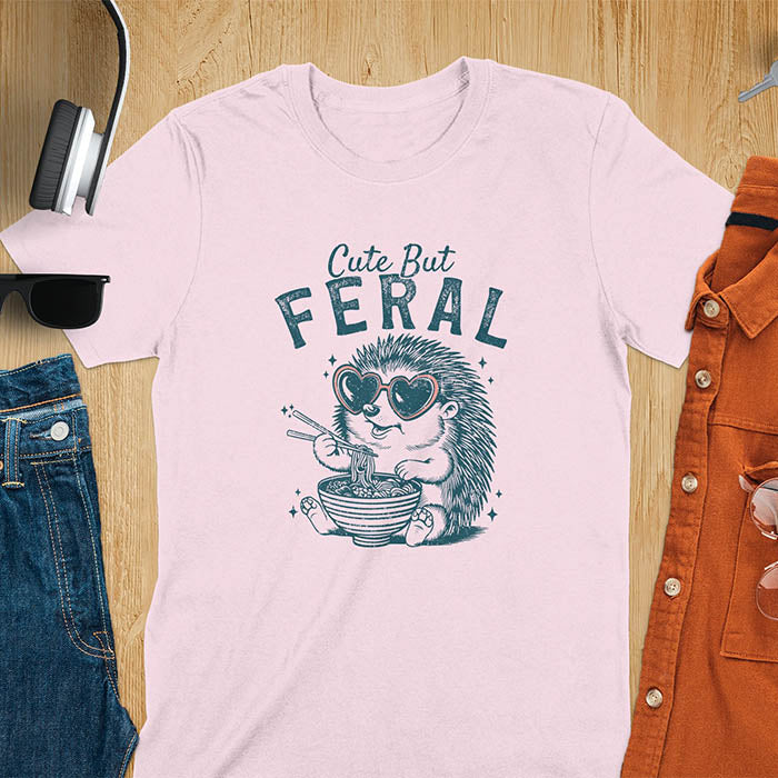 A t-shirt with a hedgehog wearing heart sunglasses and eating ramen with chopsticks. Text: "Cute But" above and "FERAL" below. This kawaiiness cuteness shows hedgehog cuteness in a weird funny shirt while ramen eating, a funny animal meme. A meme graphic tee and funny unhinge shirt, a great animal person gift and pet owner gift. A gen z meme with retro aesthetics tee vibes, a gift for mom or hers option, not quite wild safari animals.