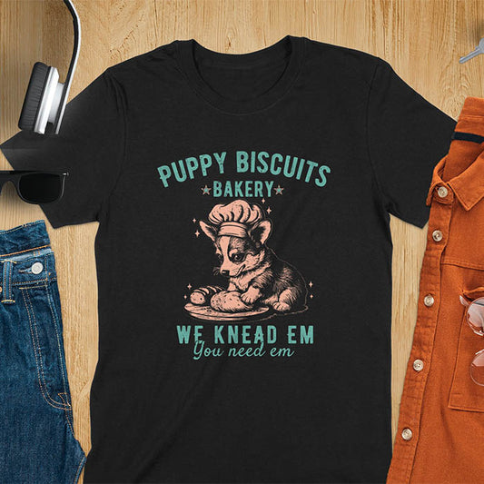 A t-shirt with a corgi in a baker's hat, with its paws on a baking surface with dough. Text: "PUPPY BISCUITS BAKERY" above "We Knead Em You Need Em". This funny baking shirt is a funny meme shirt displaying corgi cuteness, a dog lover gift and gift for dog mom. An animal lovers tshirt and dog mom shirt, a corgi dad t-shirt or pet dog owner gift. One of the funny bakers gifts, a gift for mom.