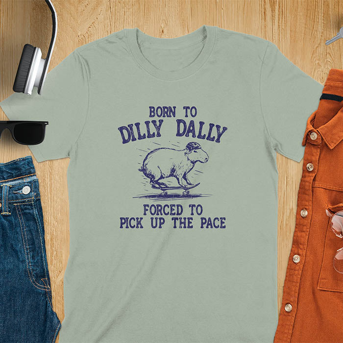 A capybara skateboarding, donning a protective helmet .Above it, the words "Born to dilly dally", while below, "Forced to pick up the pace". Rendered in a charming vintage font with a faded look.