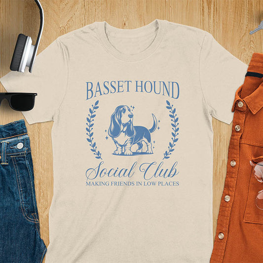 A t-shirt featuring a vintage-style basset hound design in muted blue. Includes Basset Hound, Social Club, Short & Sweet Forever text, laurel wreaths, EST 2024, and a centered dog illustration for a timeless, elegant look.