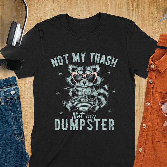 A t-shirt featuring a raccoon wearing pink heart-shaped sunglasses, sitting with chopsticks and eating ramen from a bowl. Small flies surround the raccoon. Text: NOT MY TRASH above and Not my DUMPSTER below in a distressed, bold font. The raccoon has a striped tail and a cartoonish appearance.