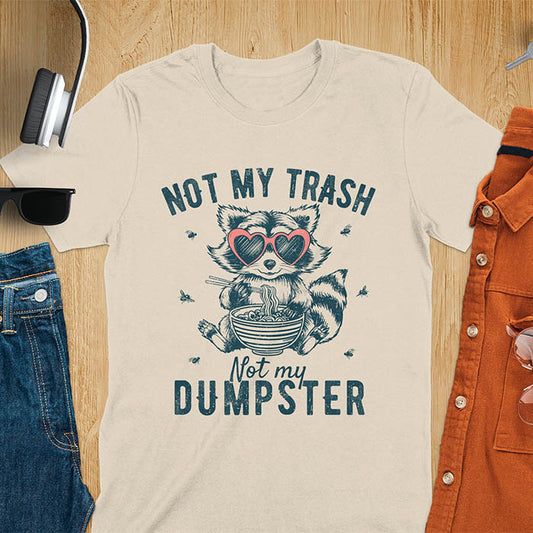A t-shirt featuring a raccoon wearing pink heart-shaped sunglasses, sitting with chopsticks and eating ramen from a bowl. Small flies surround the raccoon. Text: NOT MY TRASH above and Not my DUMPSTER below in a distressed, bold font. The raccoon has a striped tail and a cartoonish appearance.