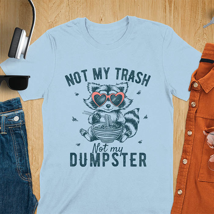 A t-shirt featuring a raccoon wearing pink heart-shaped sunglasses, sitting with chopsticks and eating ramen from a bowl. Small flies surround the raccoon. Text: NOT MY TRASH above and Not my DUMPSTER below in a distressed, bold font. The raccoon has a striped tail and a cartoonish appearance.