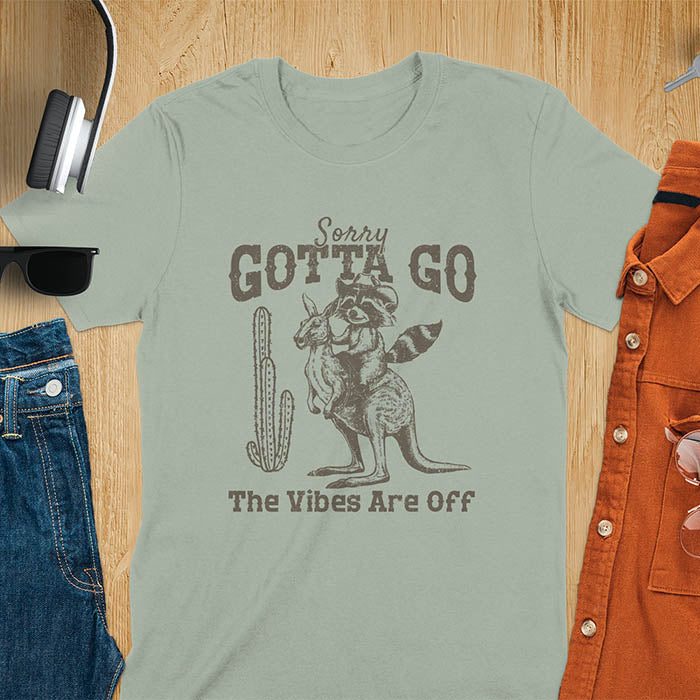 A t-shirt with a raccoon wearing a cowboy hat riding a kangaroo. Text: Sorry GOTTA GO above and The Vibes Are Off below. This humorous tee-shirt features a funny gen z meme and sarcastic meme shirt, a weird graphics shirt. A funny retro graphic and vintage graphic tee with rodeo cowboy elements. A silly shirt or funny unhinged shirt, an animal person gift or kangaroo gifts. A raccoon t shirt or mom gift alternative.