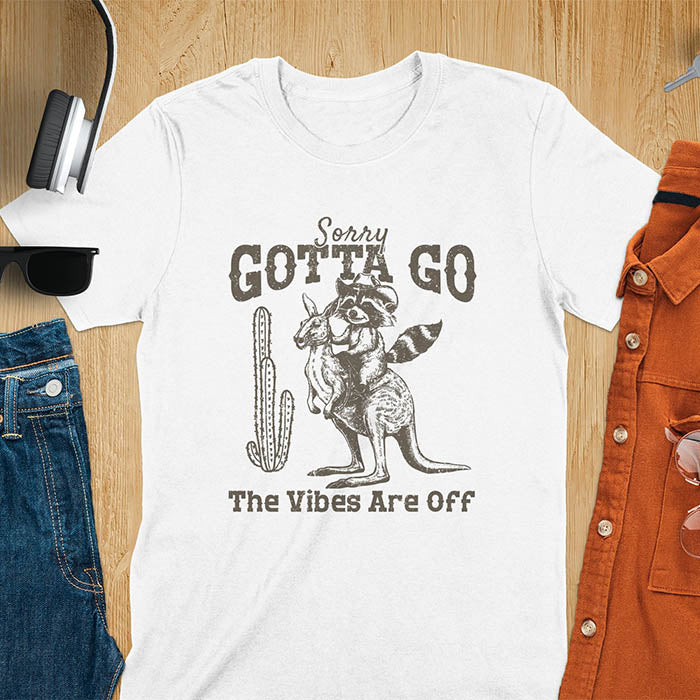 A t-shirt with a raccoon wearing a cowboy hat riding a kangaroo. Text: Sorry GOTTA GO above and The Vibes Are Off below. This humorous tee-shirt features a funny gen z meme and sarcastic meme shirt, a weird graphics shirt. A funny retro graphic and vintage graphic tee with rodeo cowboy elements. A silly shirt or funny unhinged shirt, an animal person gift or kangaroo gifts. A raccoon t shirt or mom gift alternative.