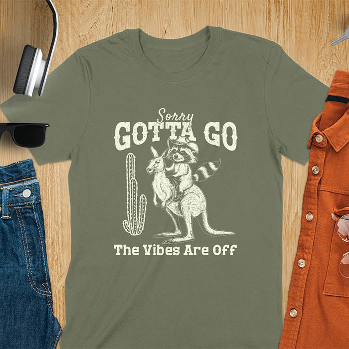 A t-shirt with a raccoon wearing a cowboy hat riding a kangaroo. Text: Sorry GOTTA GO above and The Vibes Are Off below. This humorous tee-shirt features a funny gen z meme and sarcastic meme shirt, a weird graphics shirt. A funny retro graphic and vintage graphic tee with rodeo cowboy elements. A silly shirt or funny unhinged shirt, an animal person gift or kangaroo gifts. A raccoon t shirt or mom gift alternative.